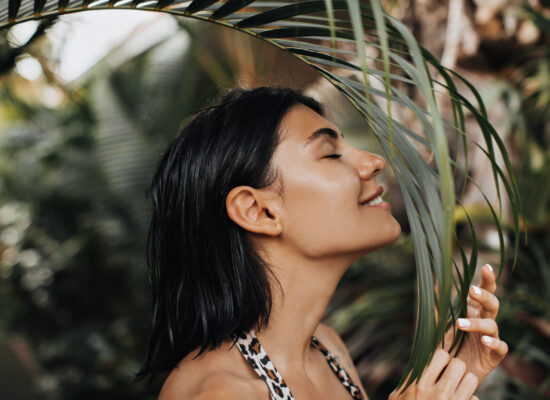 Joyful woman sniffing palm tree with closed eyes. Outdoor shot of beautiful tanned girl enjoying vacation.